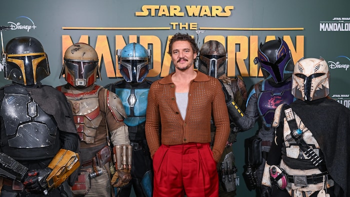 Pedro Pascal pose devant une affiche de Star Wars avec des figurines de personnages à ses côtés.