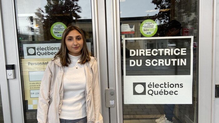 Une jeune femme debout devant une porte vitrée sur laquelle est indiqué élections Québec.