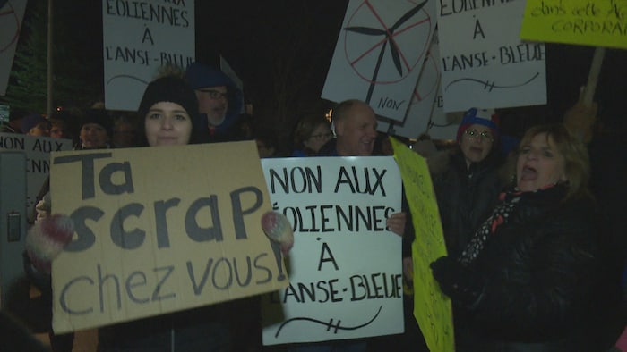 Une foule de gens qui vocifèrent et tiennent des pancartes, dont une sur laquelle est écrit « Ta scrap chez vous ».