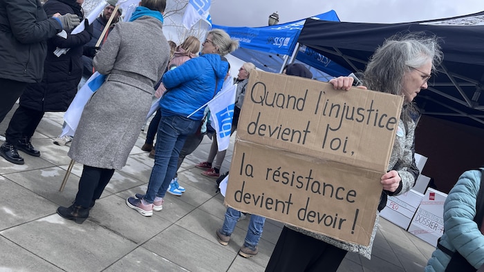  « Quand l'injustice devient loi, la résistance devient devoir ! ». Elle se trouve dans un rassemblement syndical avec des chapiteaux et d'autres manifestants.