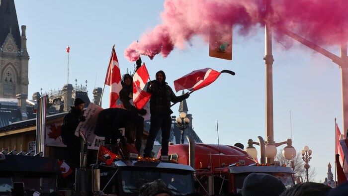 Des manifestants debout sur un camion brandissent des drapeaux et une grenade fumigène devant le parlement canadien.