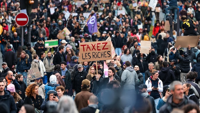Un manifestant se tient dans une foule avec une pancarte sur laquelle il est écrit « Taxez les riches ».