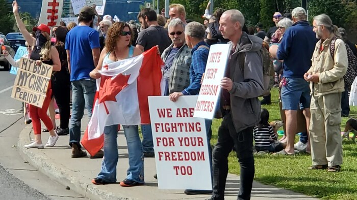 Una veintena de personas en una calle, en un día soleado, con pancartas y una bandera canadiense.