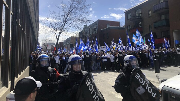 Des policiers en tenue antiémeute sont debout devant plusieurs manifestants brandissant des drapeaux du Québec dans une rue de Montréal.