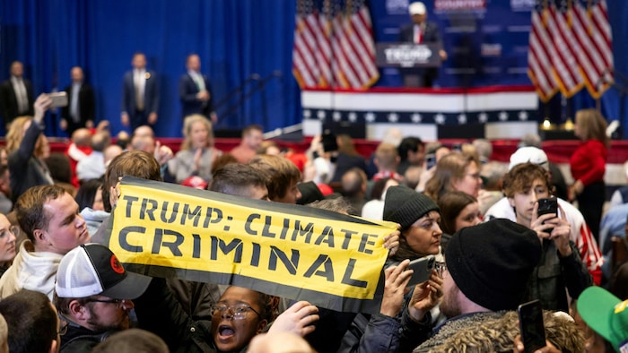 Au milieu d'une foule, une manifestante tient une banderole où l'on peut lire «Trump : criminel du climat». Loin derrière elle, Donald Trump est sur l'estrade.