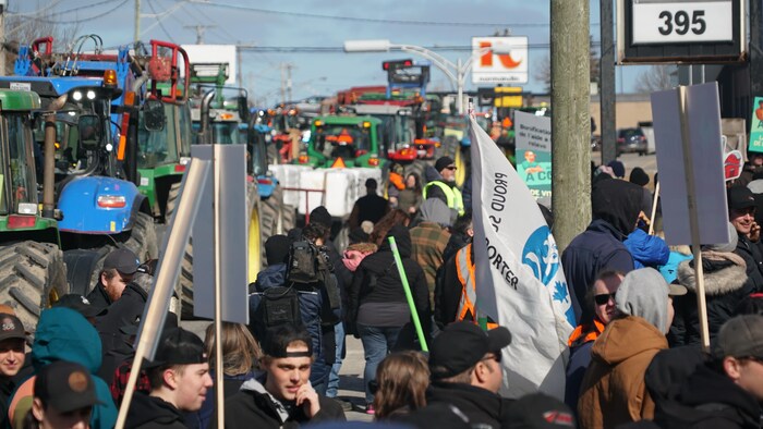 Des dizaines de personnes rassemblées avec des pancartes devant une file de tracteurs dans la rue. 
