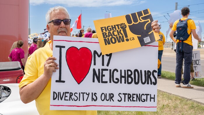 Un homme portant un polo jaune tient devant lui une grande pancarte blanche écrite à la main qui affiche un grand cœur rouge et l'inscription « I (love) MY NEIGHBOURS DIVERSITY IS OUR STRENGTH ».