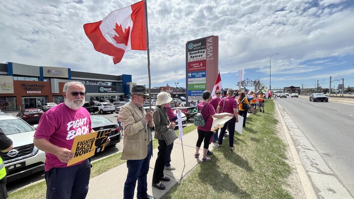 Un groupe de manifestants est rassemblé le long d'un trottoir bordant une grande artère routière, devant un centre commercial avec au premier plan, un homme barbu qui tient une pancarte jaune indiquant « FIGHT BACK NOW.ca ».