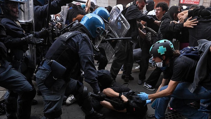 Des policiers antiémeute, portant des casques rigides, des boucliers et des matraques, chargent des manifestants et en immobilisent un au sol, qui se protège la tête.