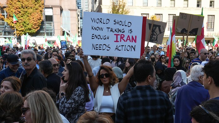 Une foule dense manifeste. Des gens brandissent des drapeaux de l'Iran et des pancartes. Trois femmes s'enlacent.
