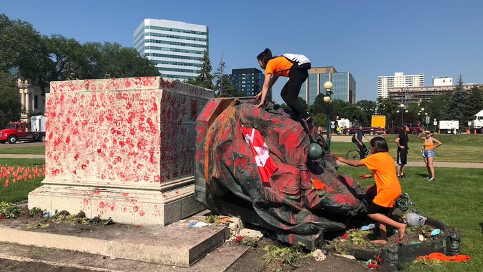 Une statue jetée au sol, couverte de peinture rouge et escaladée par des personnes portant des chandails orange.