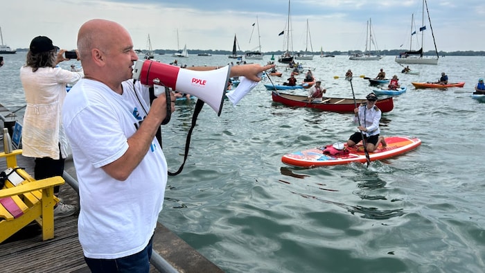 Norm Di Pasquale avec un microphone devant des gens réunis en canoë, en kayak et en bateau.