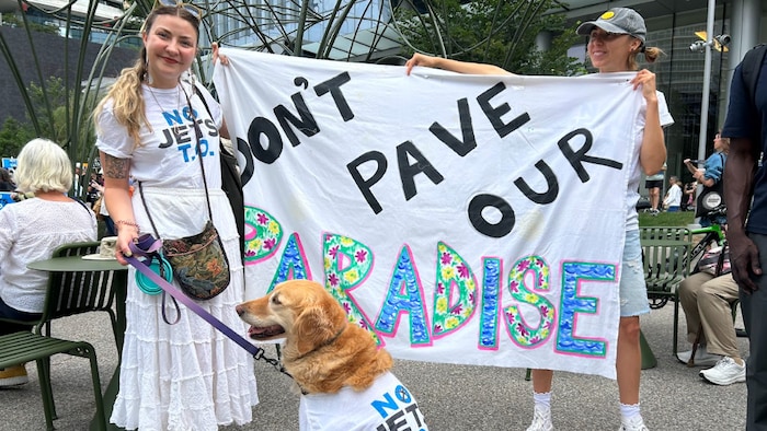 Larissa Watson et sa chienne Daisy portant toutes deux un chandail au logo de NoJetsTO devant une banderole où on peut lire « Ne pavez pas notre coin de paradis ».