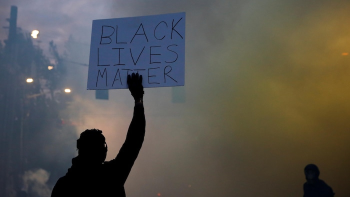 Un homme, de dos, brandit une pancarte sur laquelle il est écrit: « Black Lives Matter » dans un nuage de gaz lacrymogène.