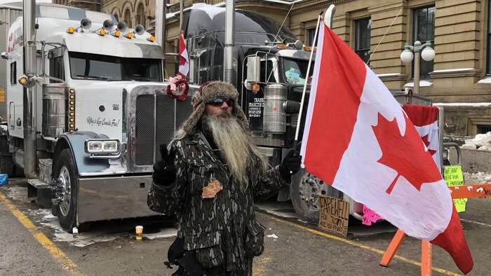 Une personne manifeste avec un drapeau canadien à la main devant des camions.