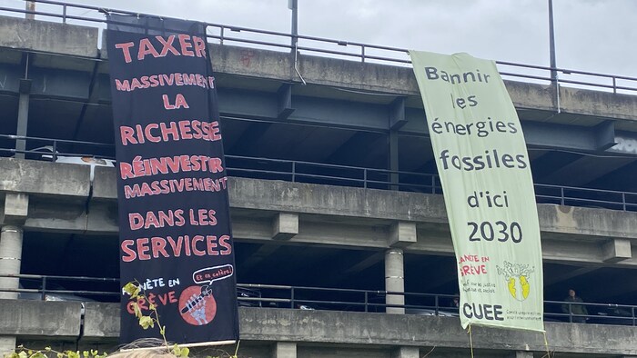 Deux immenses banderoles qui pendent d'un stationnement à étages sur lesquelles est inscrit : « Taxer massivement la richesse, réinvestir massivement dans les services » et « Bannir les énergies fossiles d'ici 2030 ».