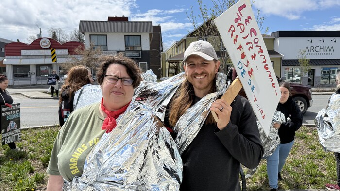 Cindy Chalifoux et Dave Longpré posent avec des couvertures de survie et une affiche dans une manifestation.