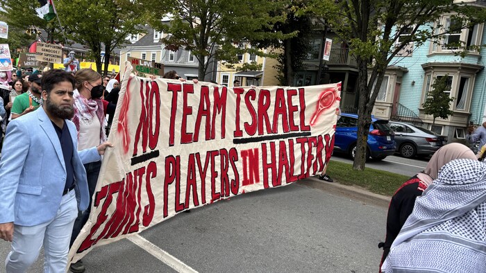 Des gens défilent dans la rue derrière une grande bannière horizontale où connected  a peint en rouge la operation   «No Team Israel Tennis Players successful  Halifax».