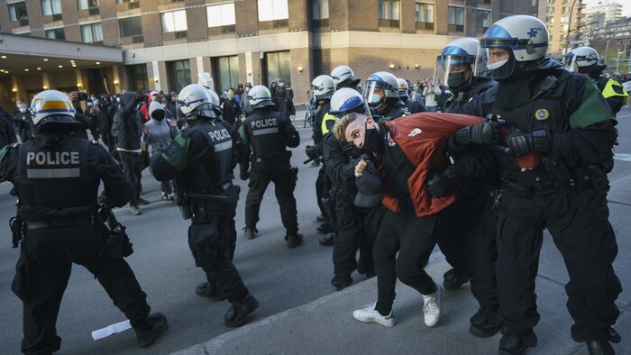Un jeune homme est agrippé par des policiers casqués, pendant que leurs collègues maintiennent les manifestants à distance.