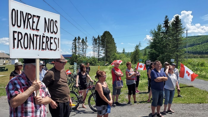 Un homme tient une pancarte «Ouvrez nos frontières!», il y a une dizaine de personnes à côté de lui et certains tiennent des drapeaux du Canada. 