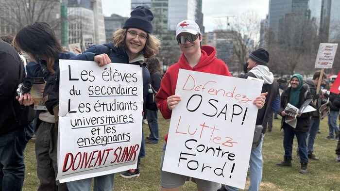 Deux jeunes tiennent des affiches disant « Luttez contre Ford » devant l'Assemblée législative à Toronto, le 24 mars 2026.