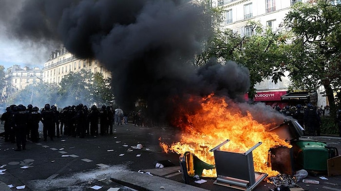 Une vingtaine de policiers montent la garde près de poubelles et de meubles qui sont dévorés par les flammes sur une rue de Paris.