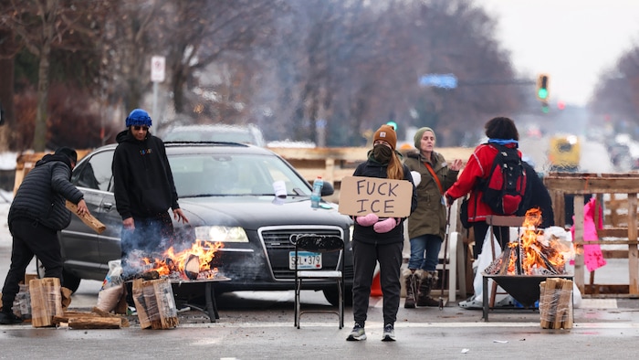 Des manifestants tiennent une pancarte et alimentent des feux de bois devant une barricade faite de bois de palettes.