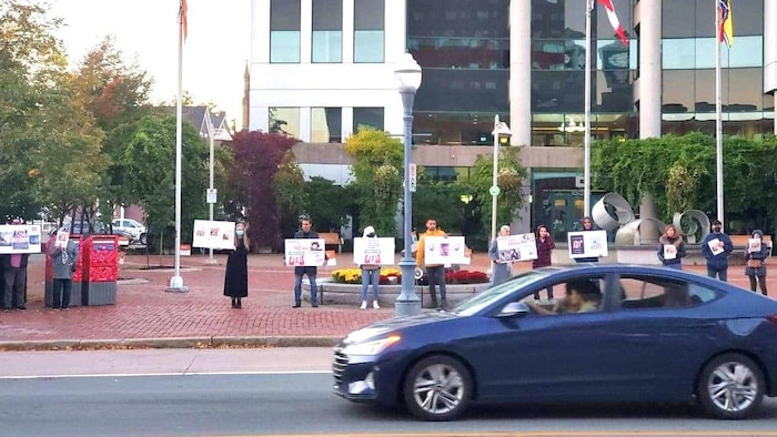 Une automobile passe devant un groupe de manifestants alignés sur le trottoir avec des pancartes.