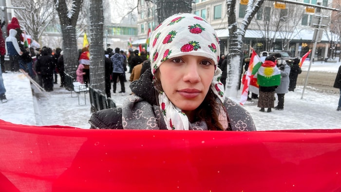 Une jeune femme portant un foulard blanc à motifs floraux roses sur la tête et une écharpe grise. Elle tient devant elle un grand drapeau kurde.