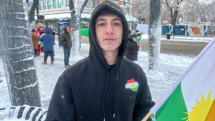 Un jeune homme portant un chandail à capuchon noir orné d'un petit logo aux couleurs du Kurdistan sur la poitrine tient un drapeau kurde fixé à un bâton de bois.