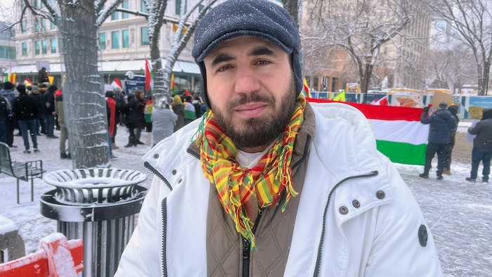 Danyar Bradost se tient sur la place de l'hôtel de ville de Calgary enneigé où l'on aperçoit, en arrière-plan, un rassemblement de personnes avec des drapeaux kurdes.