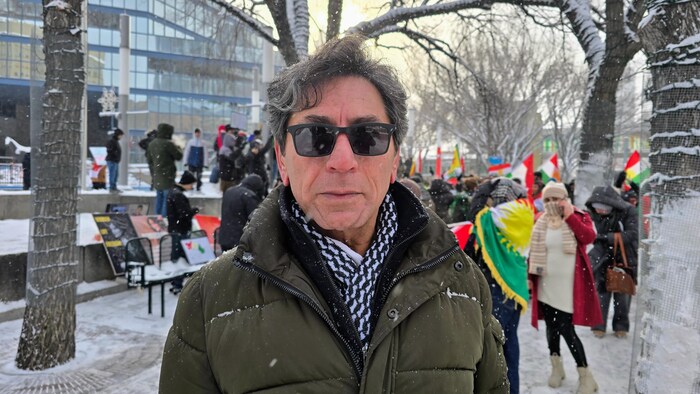 Un homme aux cheveux grisonnants, portant des lunettes de soleil noires, un manteau d'hiver et une écharpe à motifs noirs et blancs, se tient devant l'hôtel de ville de Calgary. À l'arrière-plan, plusieurs personnes sont rassemblées pour une manifestation, certaines brandissant des drapeaux kurdes.