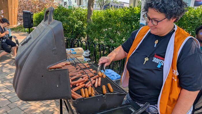 Une dame apprête des saucisses sur un barbecue en souriant.