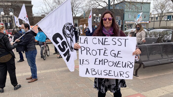 Une femme tient une affiche disant «La CNESST protège les employeurs abuseurs».