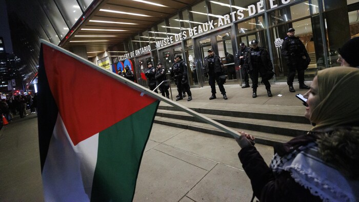 Une personne tenant un drapeau de la Palestine devant des policiers.