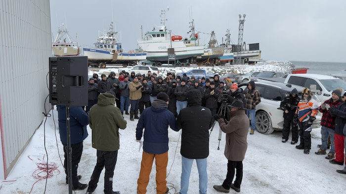 Un groupe de cinq personnes s'adresse à des gens. Au loin, des bateaux de pêche sont sur le quai pour l'hiver.