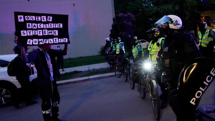 Au coin des rues Pascal et Lapierre, dans Montréal-Nord, un manifestant brandit une pancarte devant une rangée de policiers. 