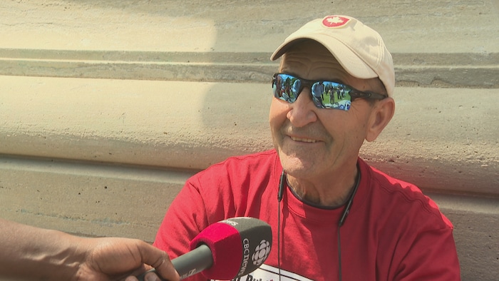 Ron Nikita portant un t-shirt rouge, des lunettes de soleil et une casquette beige ornée d’une feuille d’érable, est interviewé à l’extérieur.