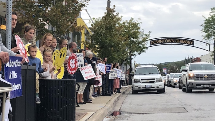 On voit à gauche une longue rangée de gens qui tiennent des pancartes contre l'avortement. À droite, on voit la rue principale avec des voitures qui circulent. Au-dessus des automobiles, on peut lire "Downtown Grimsby".