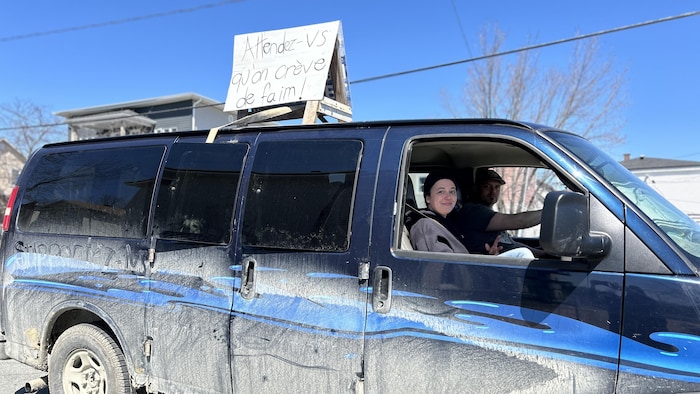 Une femme et un homme circulent dans une voiture où il est écrit « Attendez-vous qu'on crève de faim! » sur le toit.