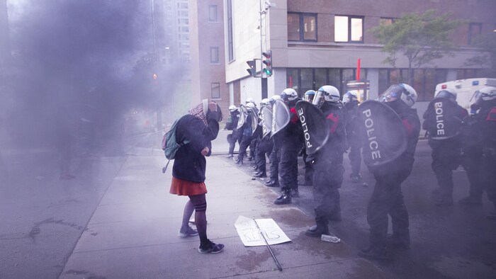 Une manifestante se couvre la tête devant une ligne de policiers en tenue antiémeute sous un nuage de gaz lacrymogène.