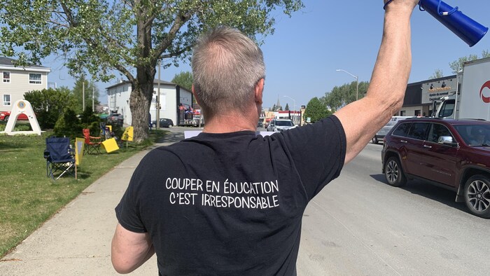 Un homme que l'on voit de dos brandit un bras et porte un chandail avec l'inscription «couper en éducation, c'est irresponsable».