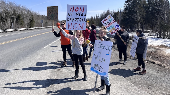 Une dizaine de personnes au bord d'une route tiennent des pancartes pour exprimer leur opposition au projet de dépotoir.