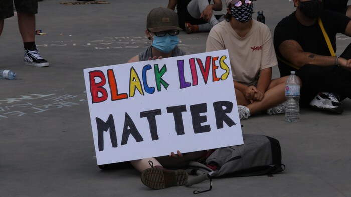 Une manifestante assise à la place Nathan Phillips de Toronto tient une pancarte sur laquelle on peut lire Black Lives Matter.