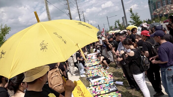 Des personnes sont rassemblées devant des affiches posées au sol où des mots sont déposés sur des post-it, et un homme au premier plan porte un parapluie jaune. 
