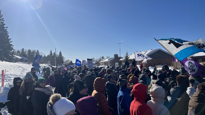 Des dizaines de personnes sont debout dans une rue résidentielle en hiver. Des drapeaux et pancartes sont visibles.