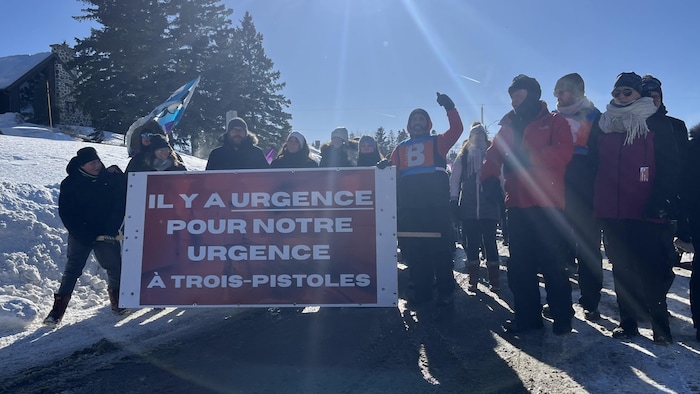 Un groupe de personnes sont debout dans une rue en hiver. Quelques personnes tiennent une grande pancarte.