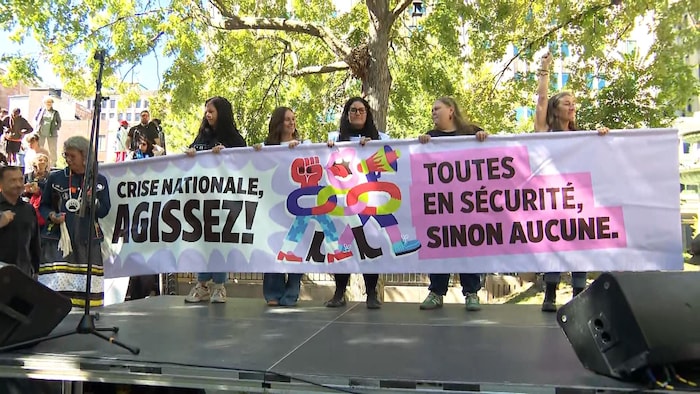 Des femmes brandissent une banderole dans un parc. Elle affiche : « Crise nationale : agissez! Toutes en sécurité, sinon aucune ».