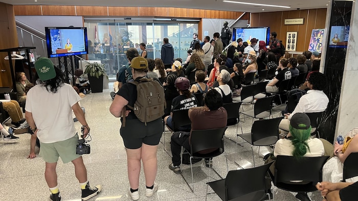 Des personnes assises dans les couloirs adjacents de la salle du conseil pour regarder le comité de dérogation sur la télévision.