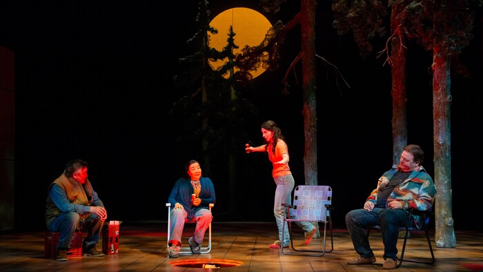 Quatre personnes assises sur une scène, un feu est devant elle, une lune à l'arrière. 